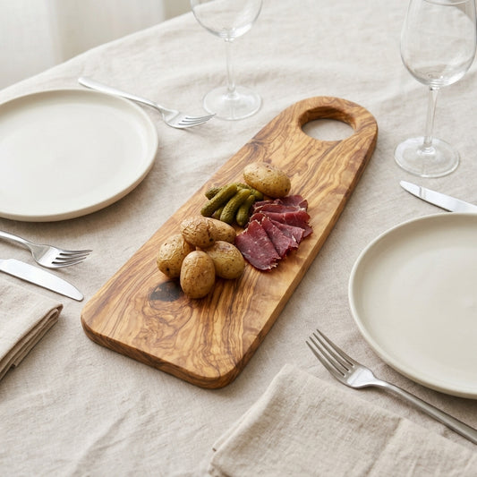 Oval olive wood board with hole handle served with potatoes, gherkins and cured meat on a laid linen table